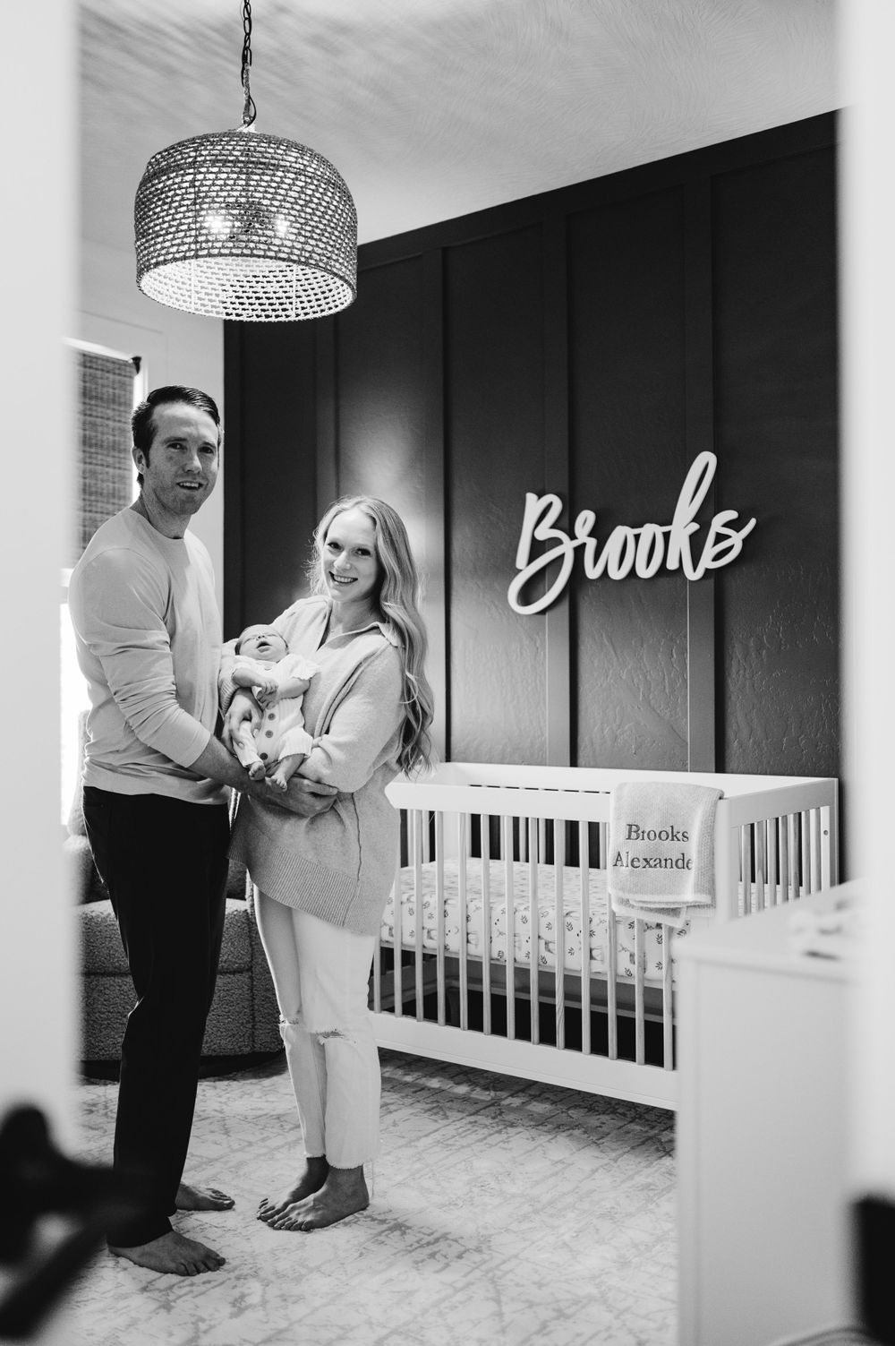Modern nursery interior with a white crib and elegant chandelier featuring the name 'Brooke' displayed on dark wall.