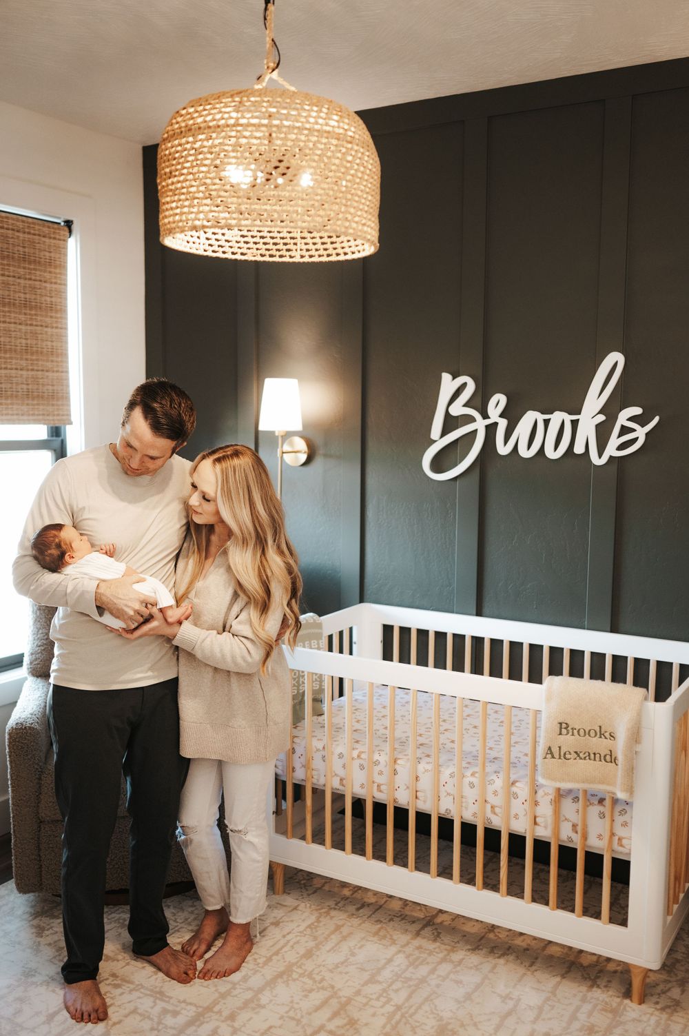 Modern nursery with woven pendant light and dark accent wall featuring white crib and illuminated name sign.