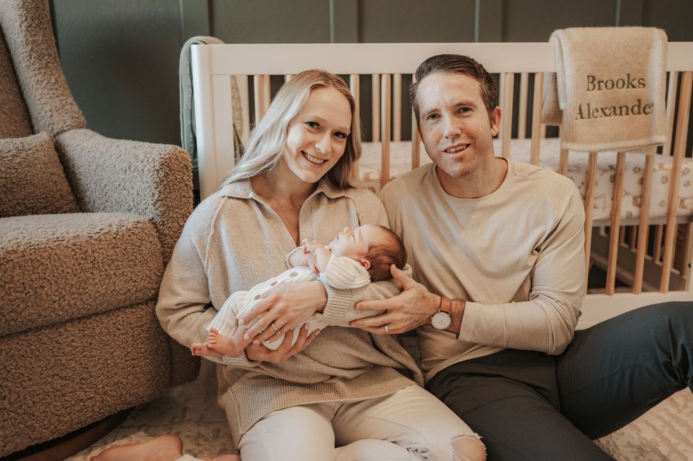 New parents relax on couch with newborn baby in nursery with crib in background.