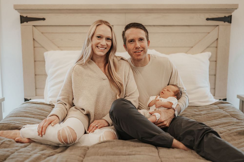 Two people in cozy beige sweaters relax together on a bed with an infant in a warm-toned indoor setting.