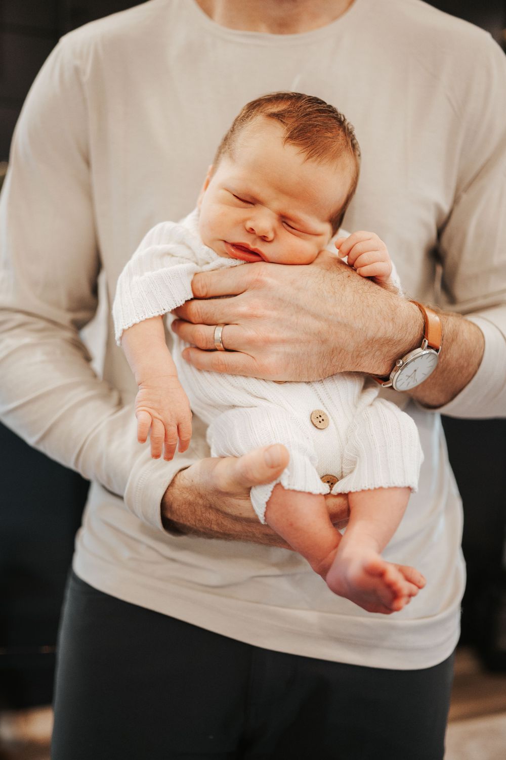A sleeping newborn in white onesie being cradled by adult hands wearing a beige sweater and wedding ring.