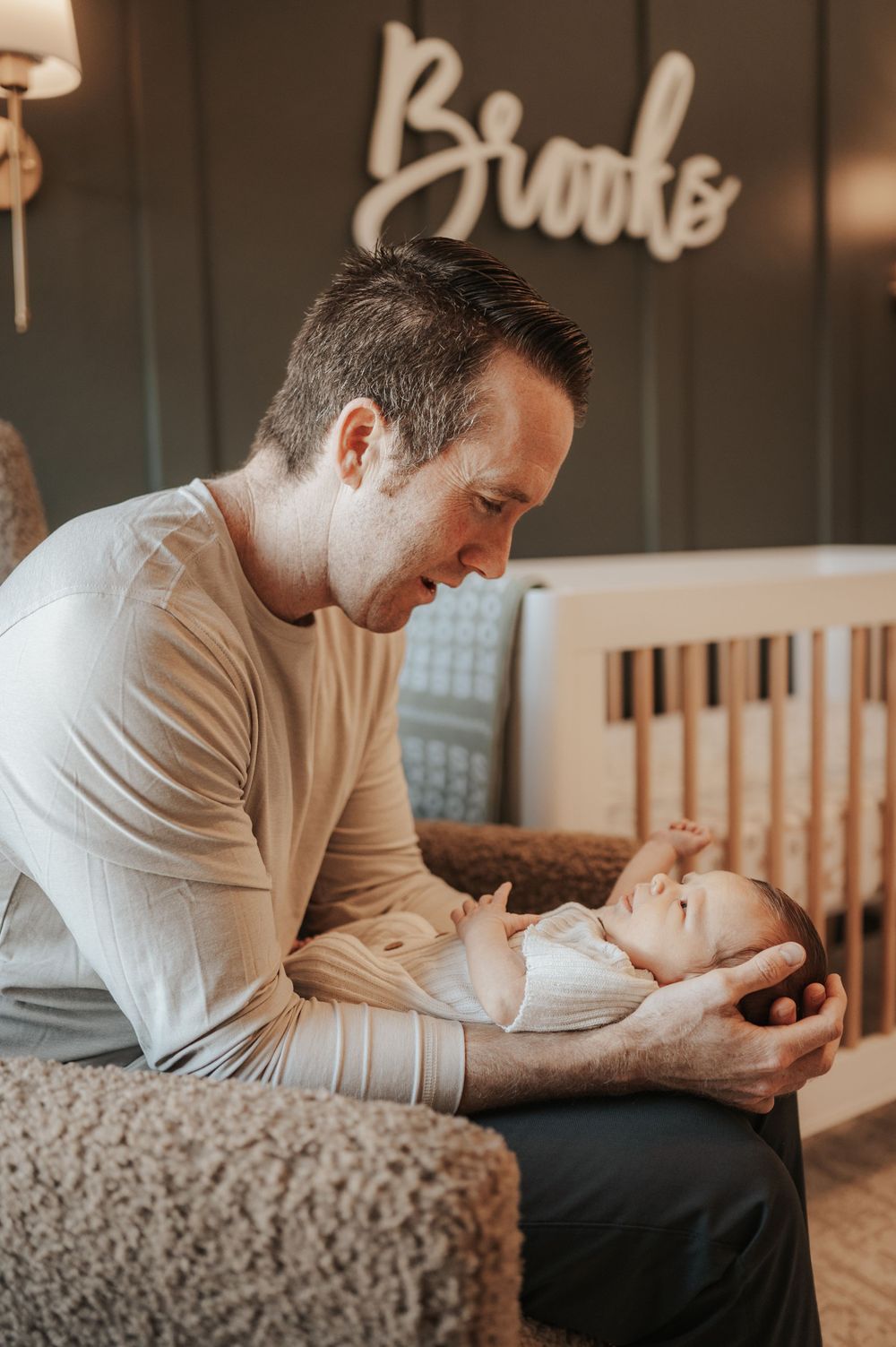 A parent gently holds and gazes at their newborn baby in a nursery decorated with the word 'Brooke'.