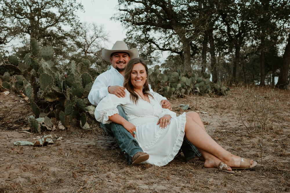 Couple relaxes together on desert ground with cactus and trees in background.
