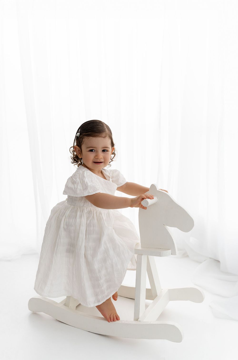 Child in white dress sits on white wooden rocking horse against light background.
