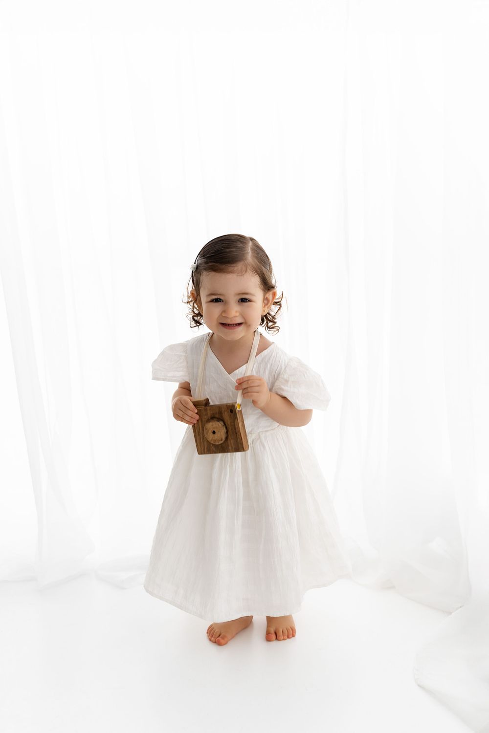 Small child in white dress holds vintage camera while standing barefoot against bright white backdrop.