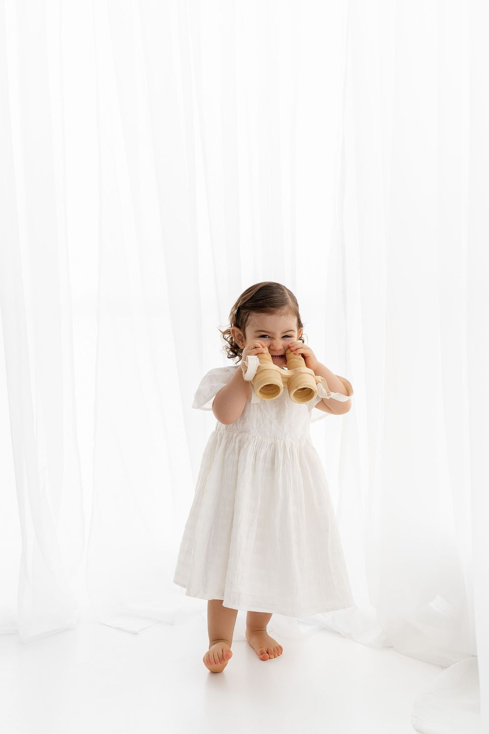 A small child in a white dress holds onto a loaf of bread while standing on a bright white backdrop.