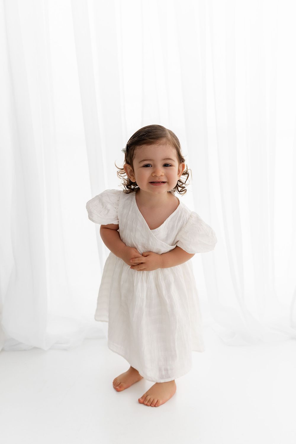 Small child in flowing white dress stands barefoot against white curtain backdrop in elegant studio setting.