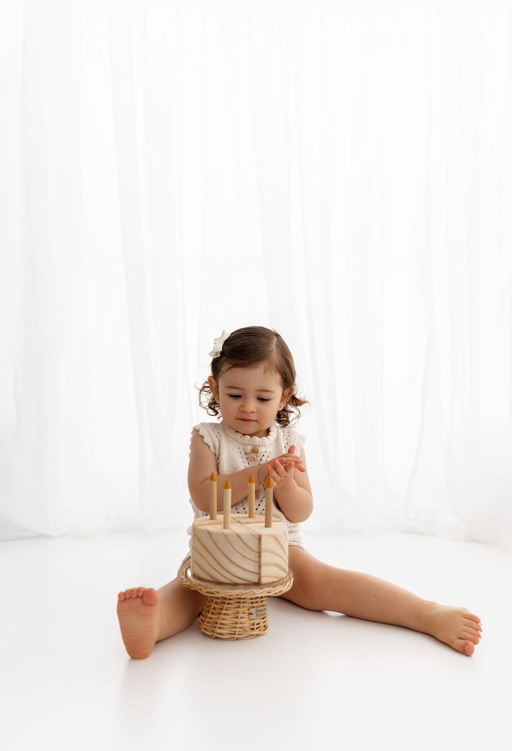 Small toddler in cream-colored outfit sits on floor with basket and birthday candles in minimalist white setting.
