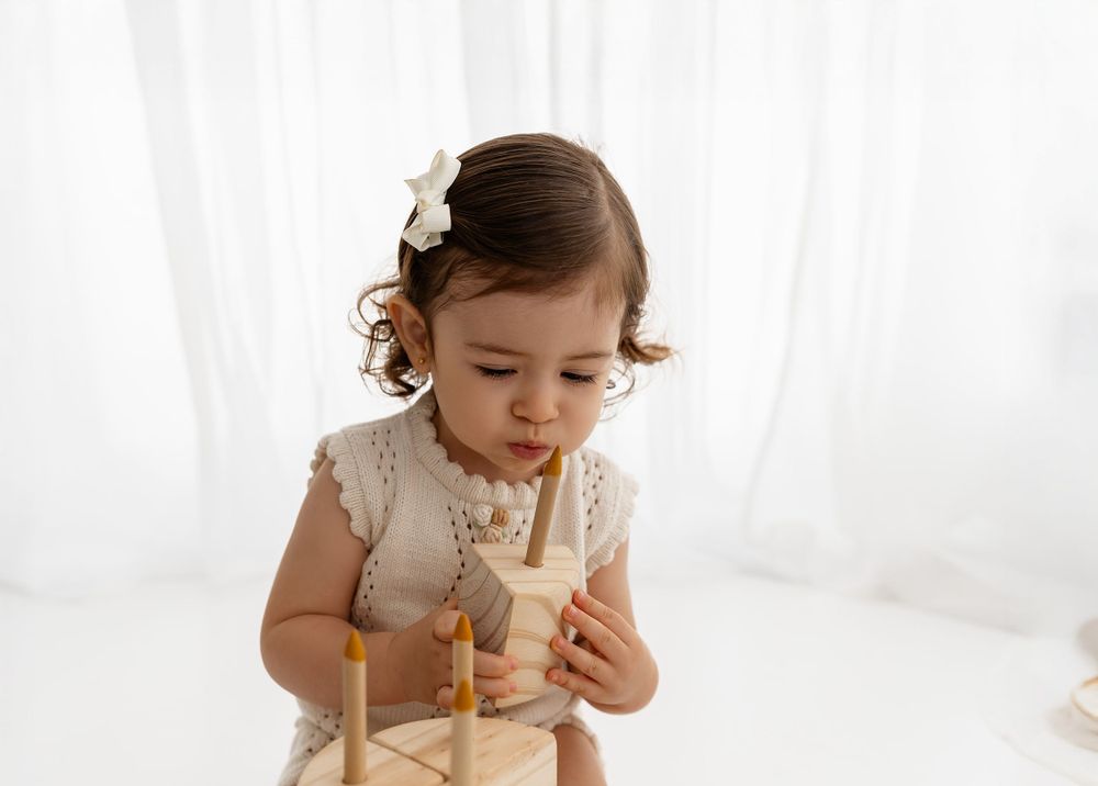 Young child in delicate lace dress thoughtfully holds a wooden toy against white curtain background.