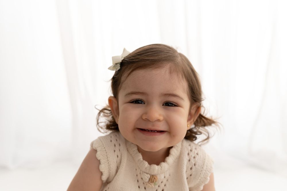 A smiling toddler in a cream-colored lace dress poses against a white backdrop in a bright studio setting.