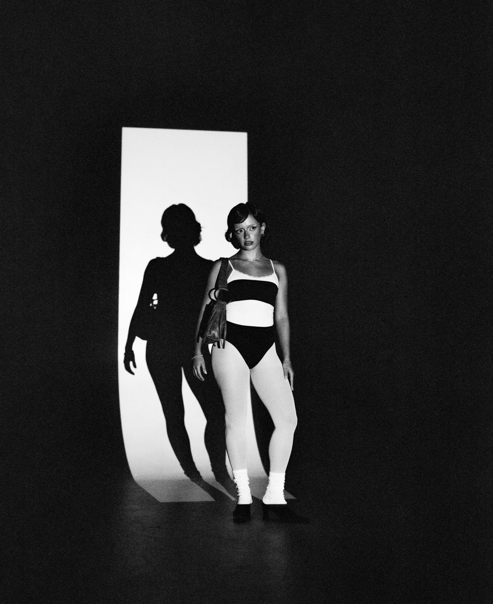 Artistic black and white dance photo showing a figure in a two-piece outfit with dramatic shadow on white backdrop.