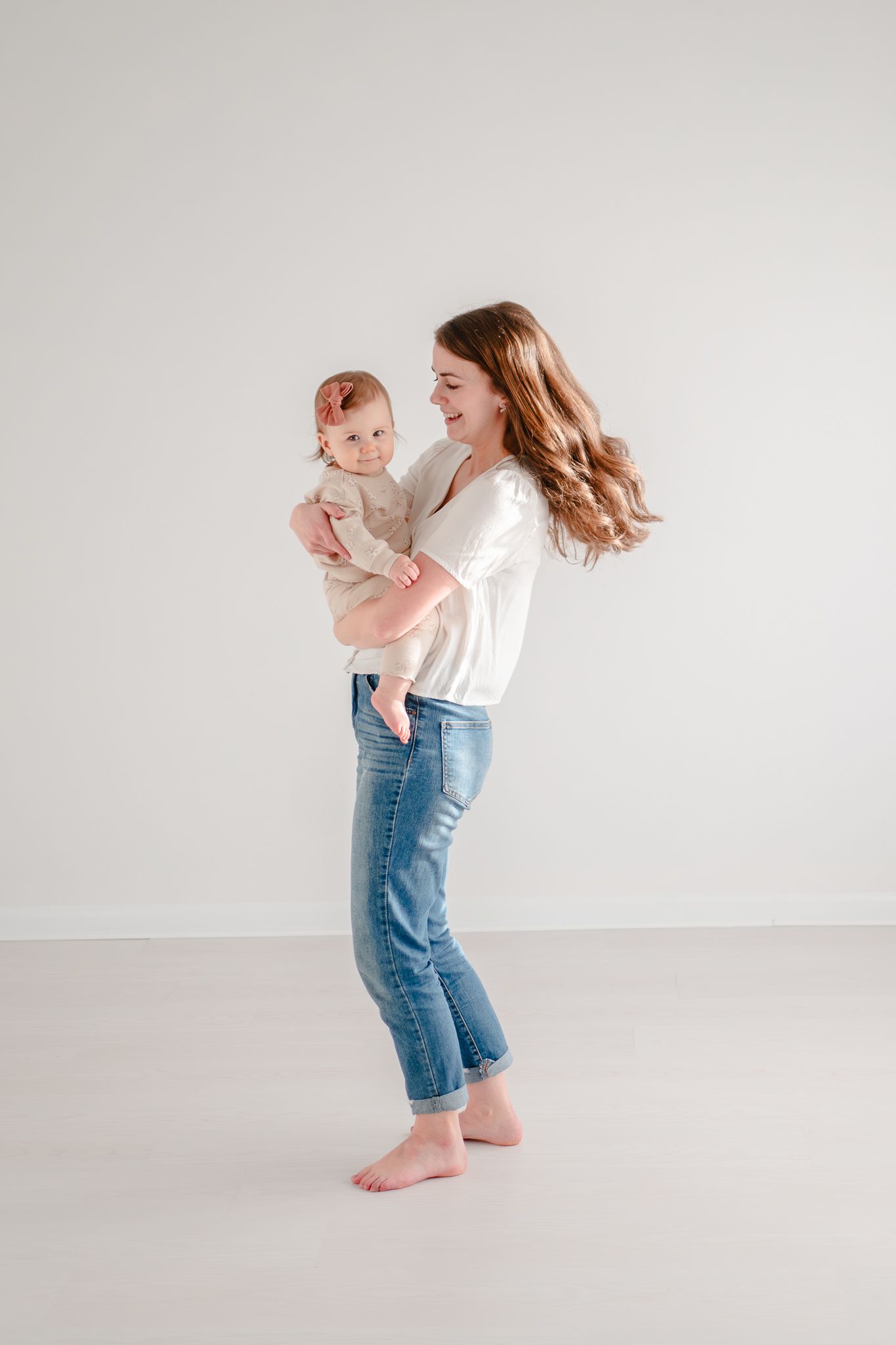 A woman in blue jeans and white top holds a baby in casual wear while dancing barefoot on a light gray background.