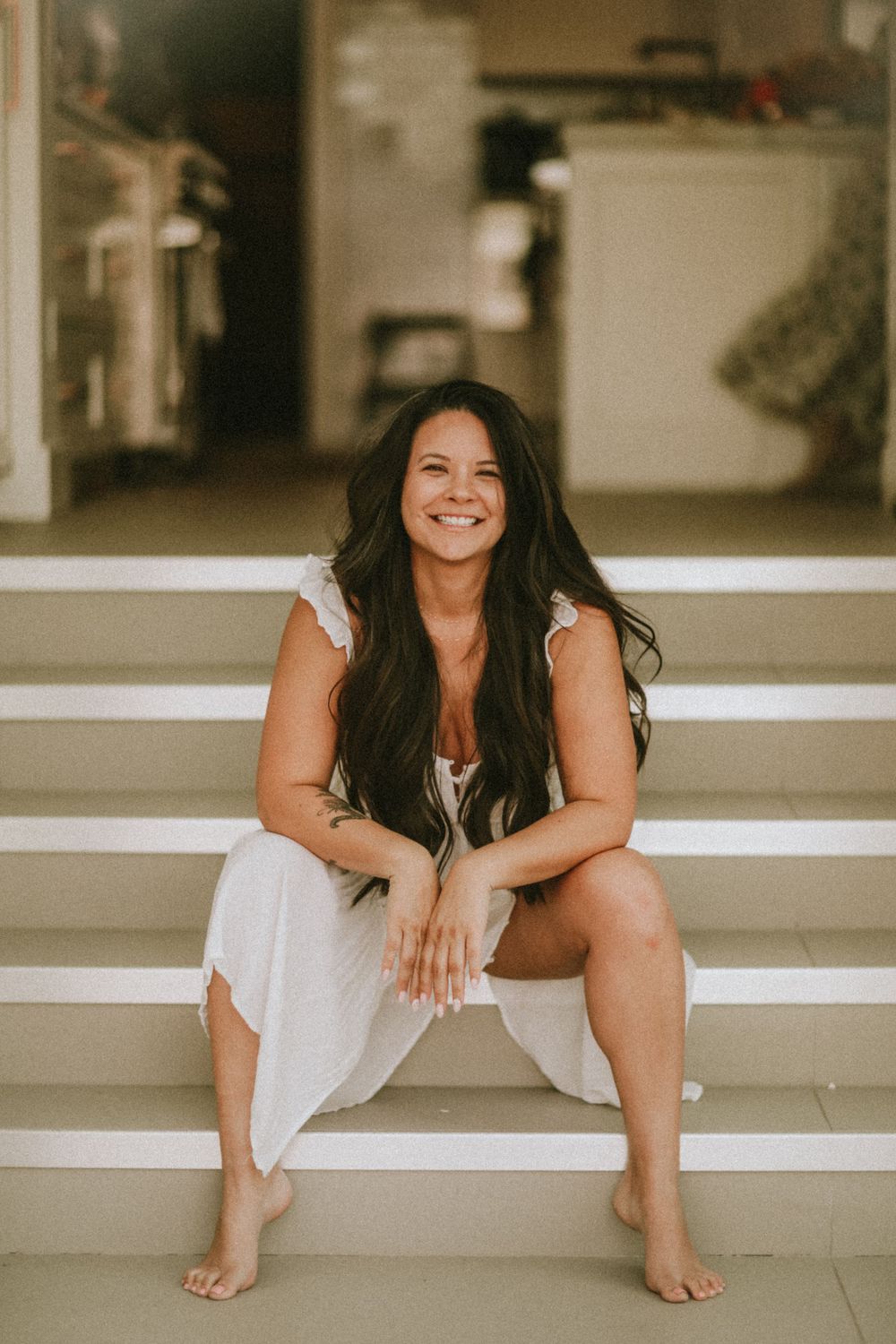 Woman in white dress sits casually on steps while smiling joyfully in natural lighting.