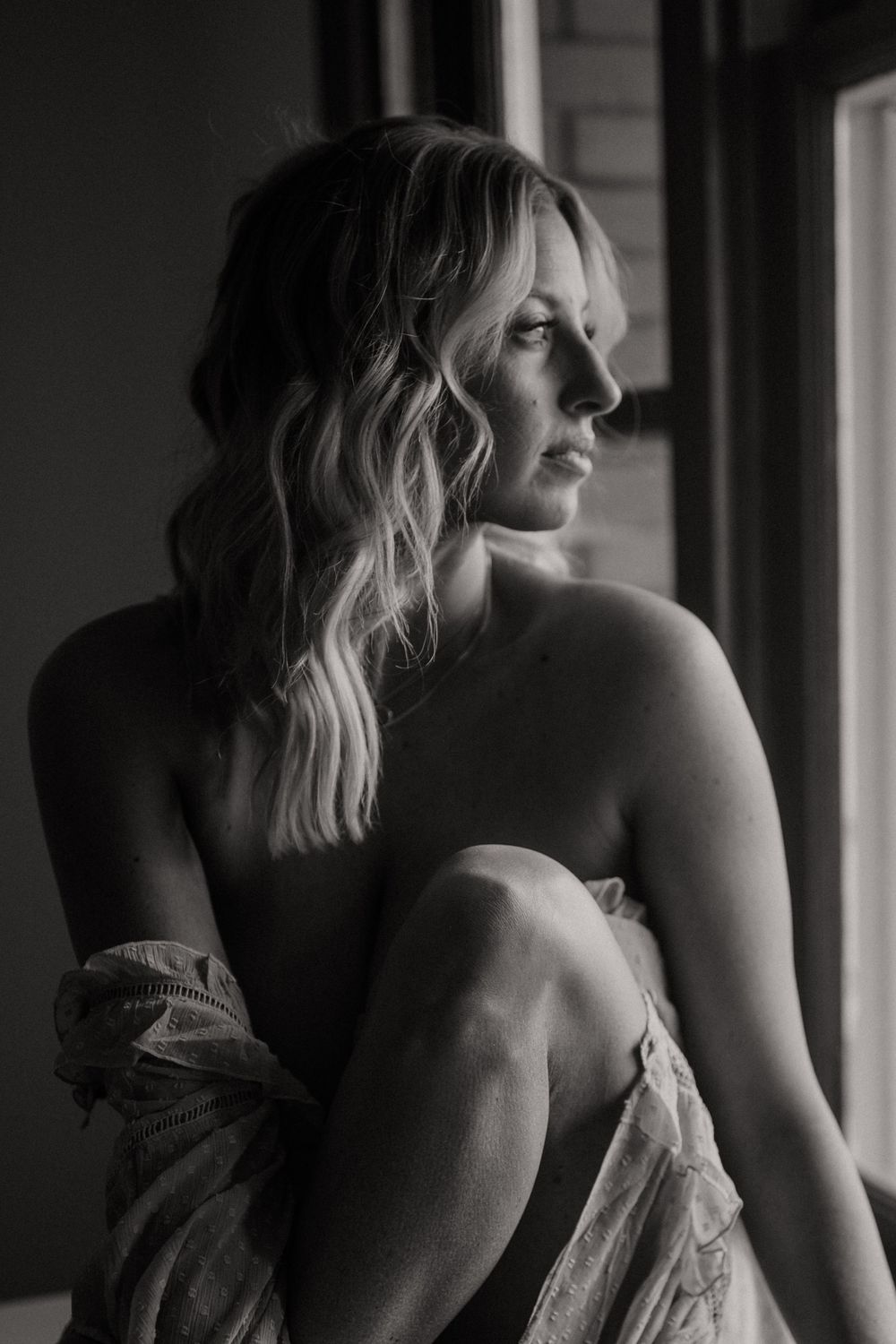 Black and white portrait of person with wavy blonde hair sitting by window in contemplative pose.