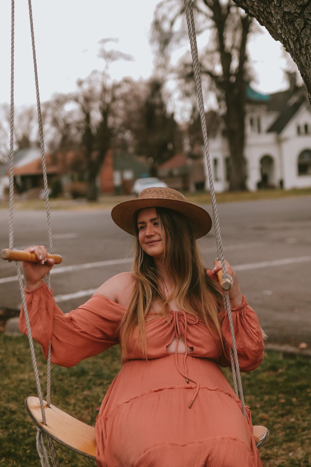 Person in a coral-colored dress and brown hat sits on a wooden rope swing in front of a house during autumn.