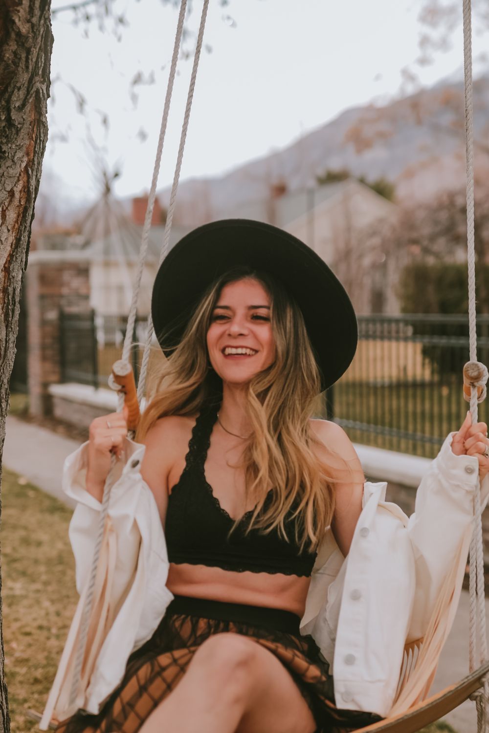 A person wearing a black bralette and white jacket sits on a wooden swing in an outdoor autumn setting.