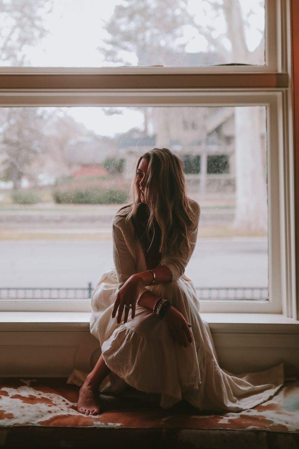 Figure in a flowing dress sits contemplatively by a window on a misty morning while soft light filters through.