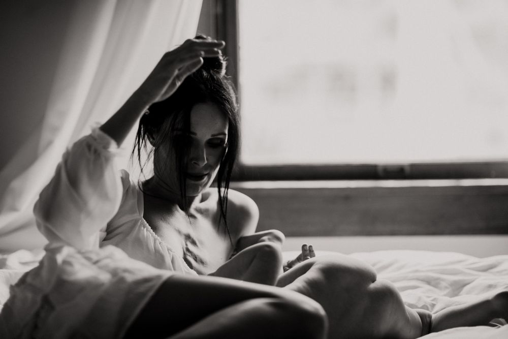 A person in white clothing reads a book by window light in a moody black and white photograph.