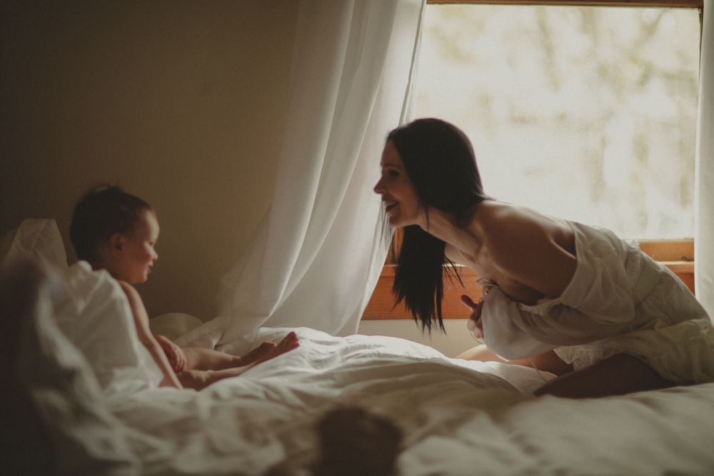 A family shares a tender moment together in a softly lit bedroom near a window with sheer curtains.