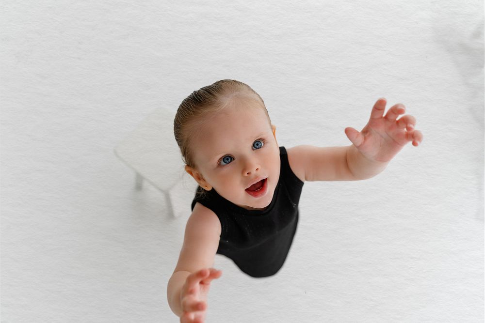 A joyful baby in a black sleeveless outfit reaching up against a white background.