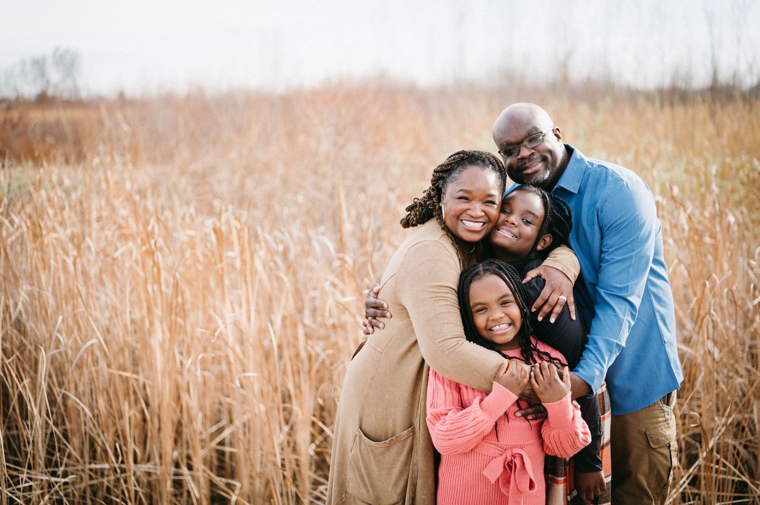 A loving family embraces while standing in a field of tall golden grass during sunset.