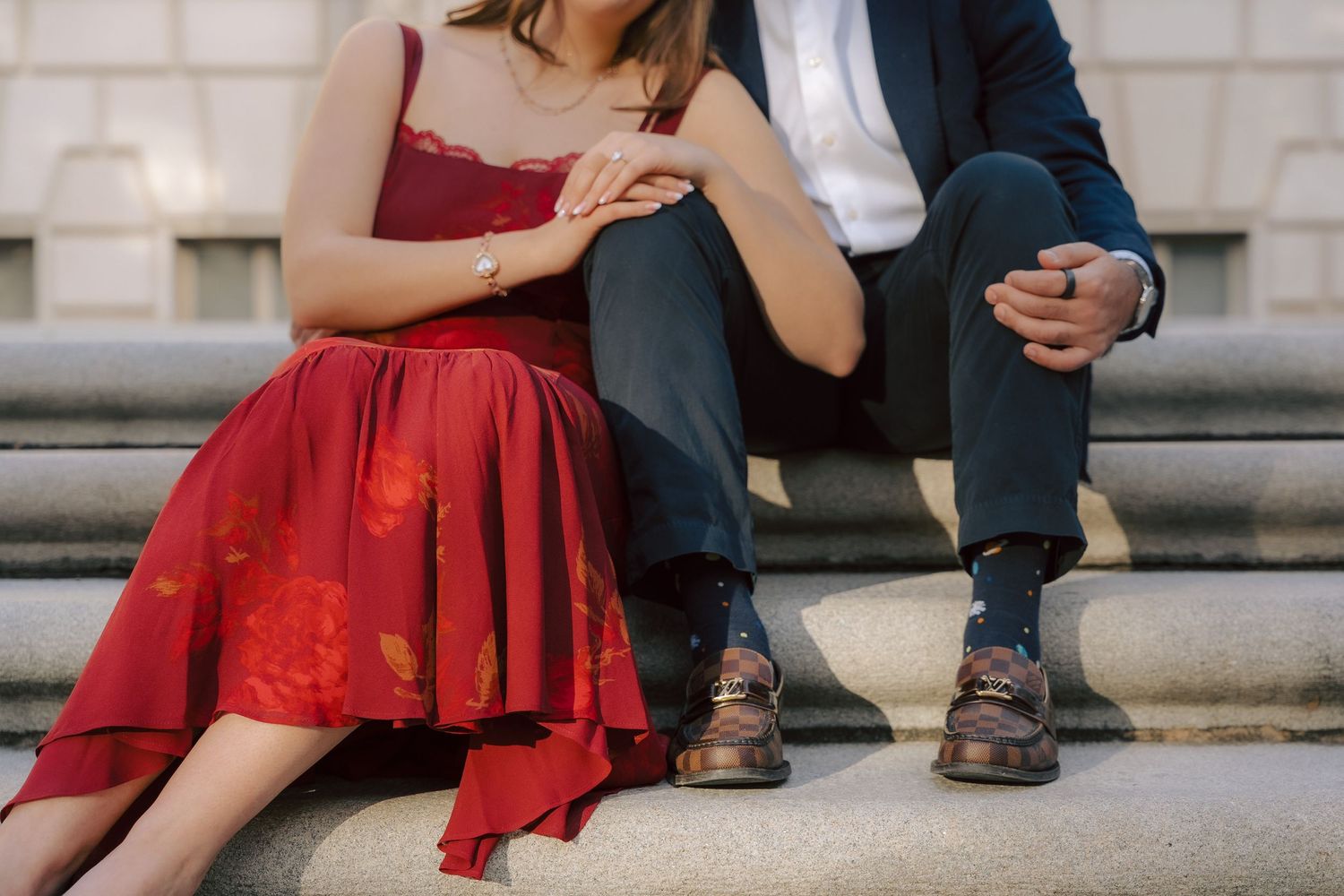 A couple sits close together on stone steps, with her in a flowing red dress and him in a dark suit.