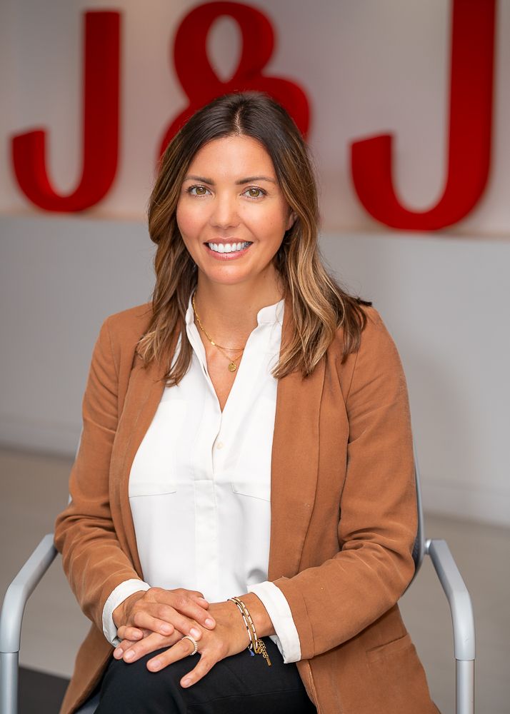 Professional woman in brown blazer and white blouse poses in office setting with red letters behind.