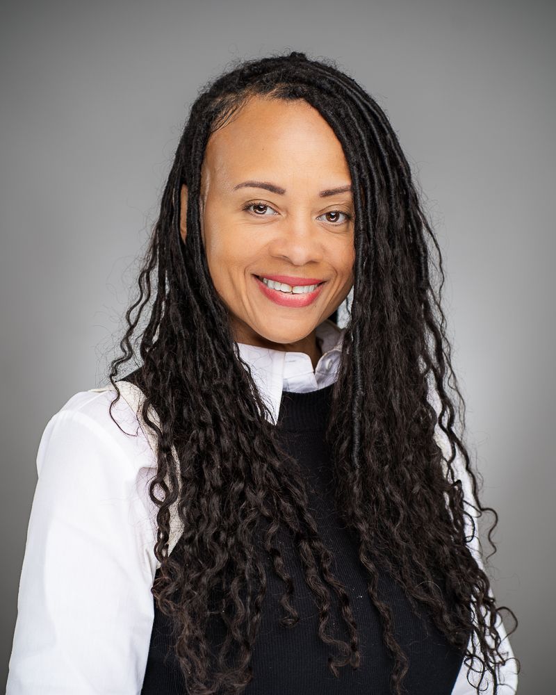 Professional headshot showing long dark braids against a gray studio backdrop.