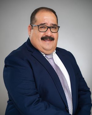Professional headshot of person in navy suit with glasses and patterned tie.
