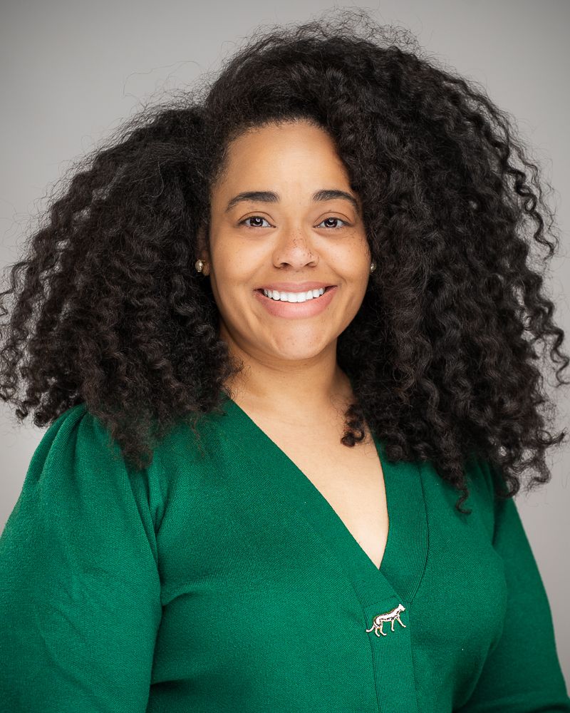 Professional portrait of someone in a green sweater against a gray background.