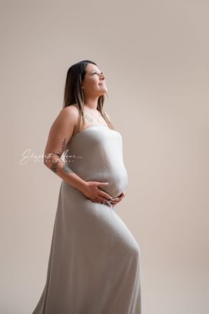A pregnant woman in a flowing white dress poses gracefully against a neutral beige backdrop during a maternity photoshoot.