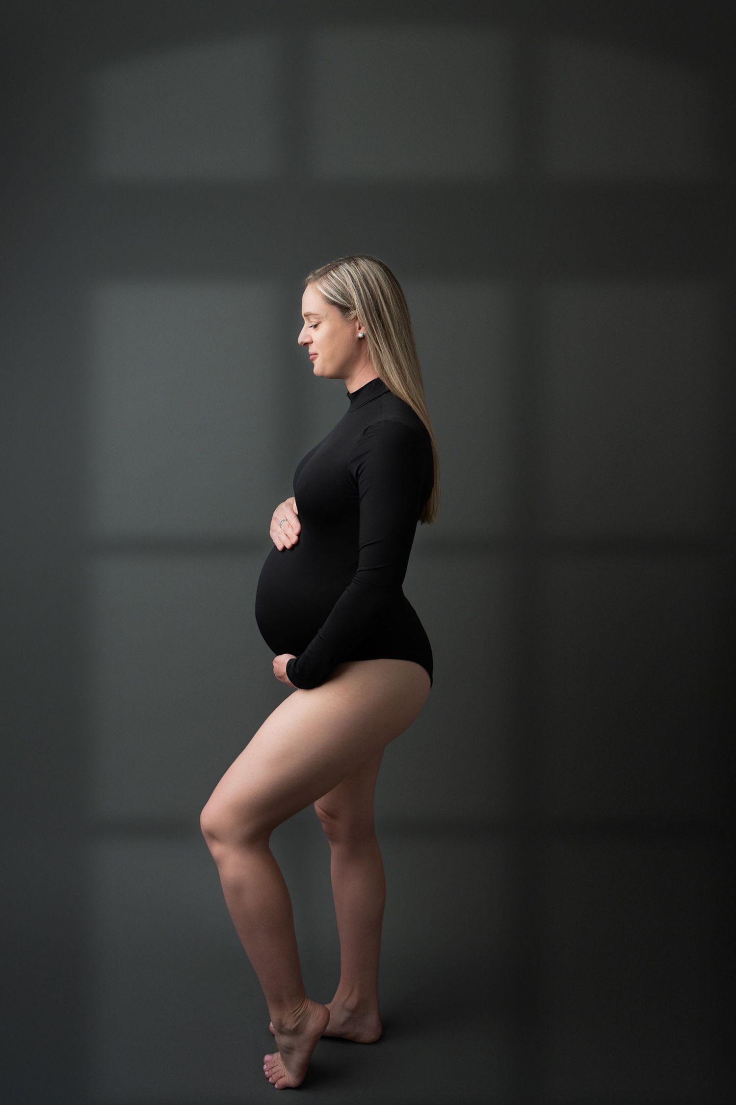 Side view of pregnant figure in black bodysuit posing elegantly against dark background in artistic maternity photo.