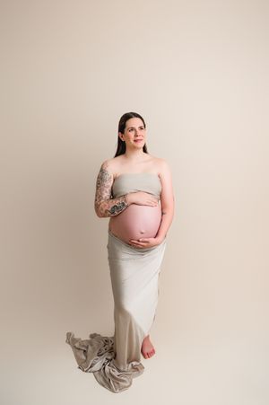 Pregnant figure in a flowing beige dress poses elegantly against neutral background, showing visible baby bump and tattoo.