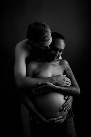Intimate black and white maternity photo featuring an embrace around a pregnant belly against dark background.