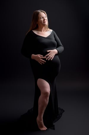 Artistic maternity photos in black dress against dark background showing silhouette and curves.