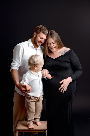 A sweet maternity photoshoot captures a family moment as they celebrate their upcoming addition against a dark backdrop.