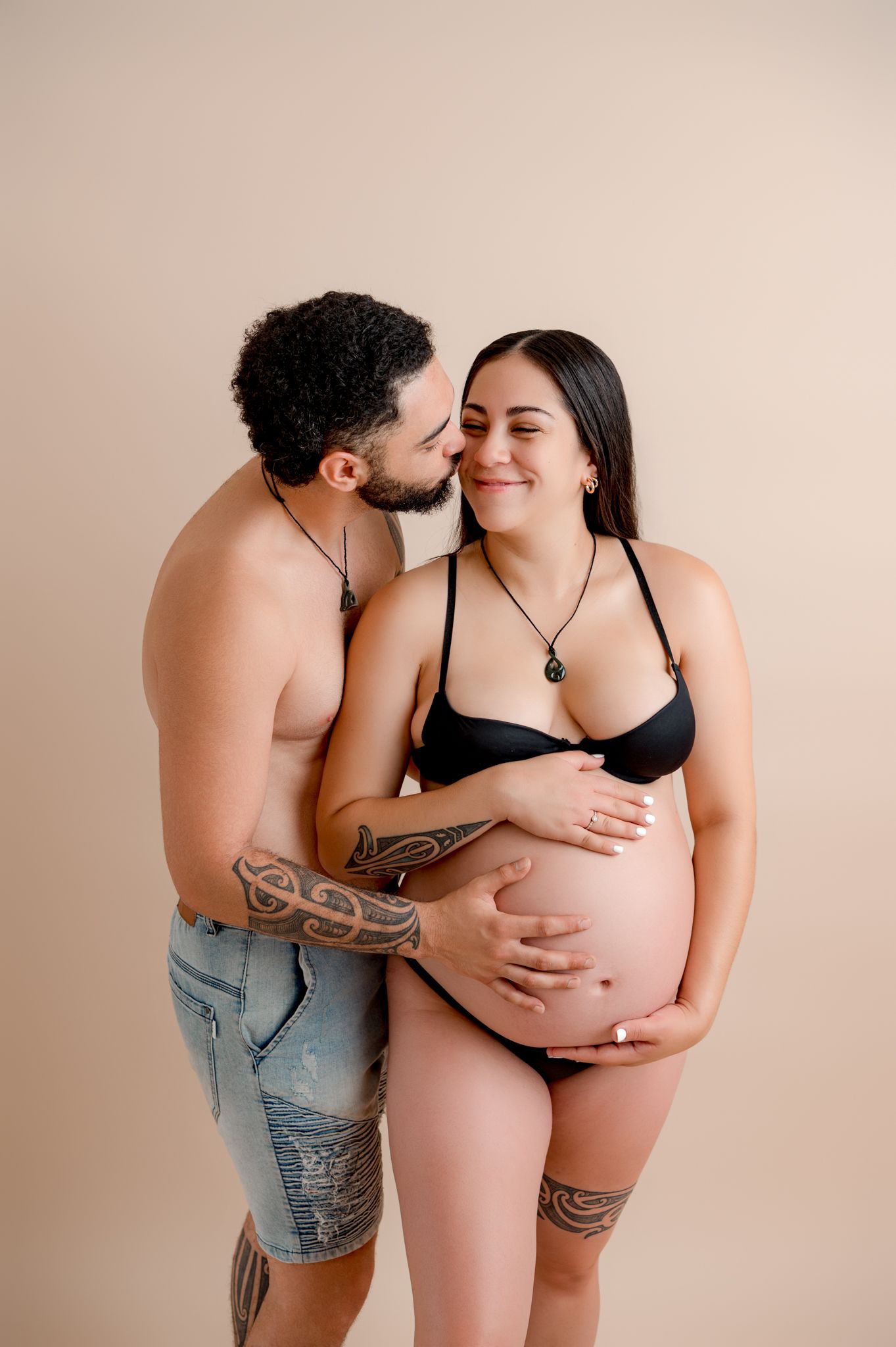 Intimate maternity photo of expecting couple embracing against beige backdrop while showing tattoos.
