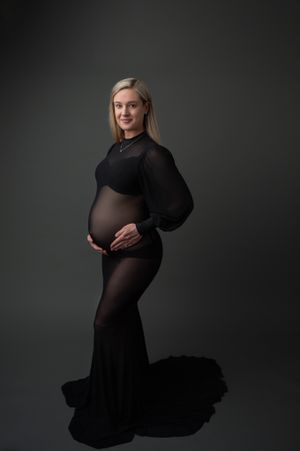 Elegant maternity portrait in black sheer gown against dark backdrop with dramatic lighting and flowing fabric.