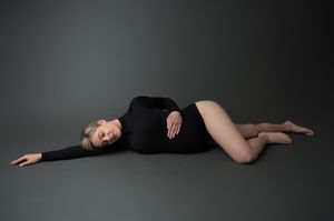 A dancer in a black leotard reclines gracefully on the floor against a dark gray studio backdrop.
