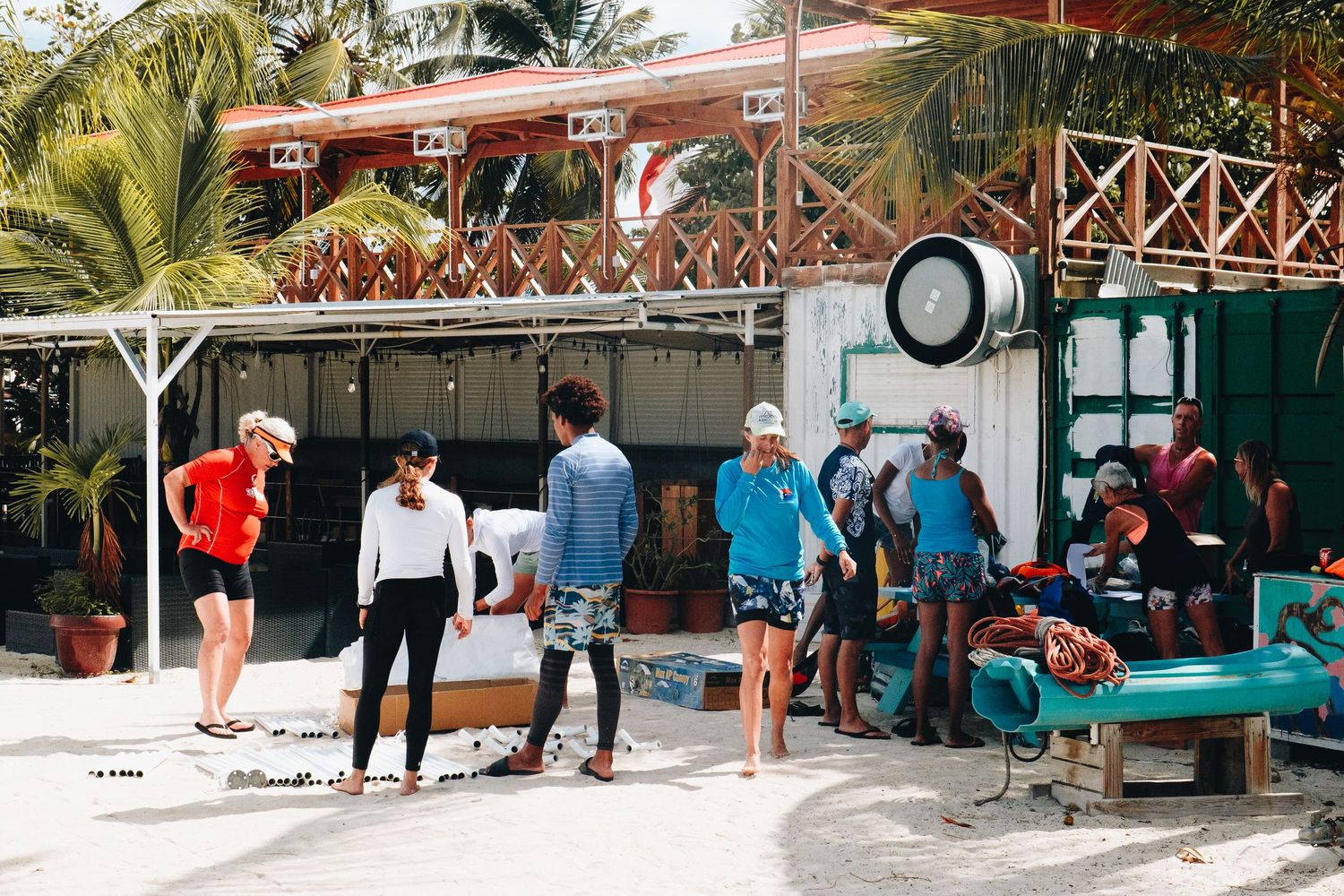 People gather outside a beach resort building with palm trees and a wooden bridge structure overhead.