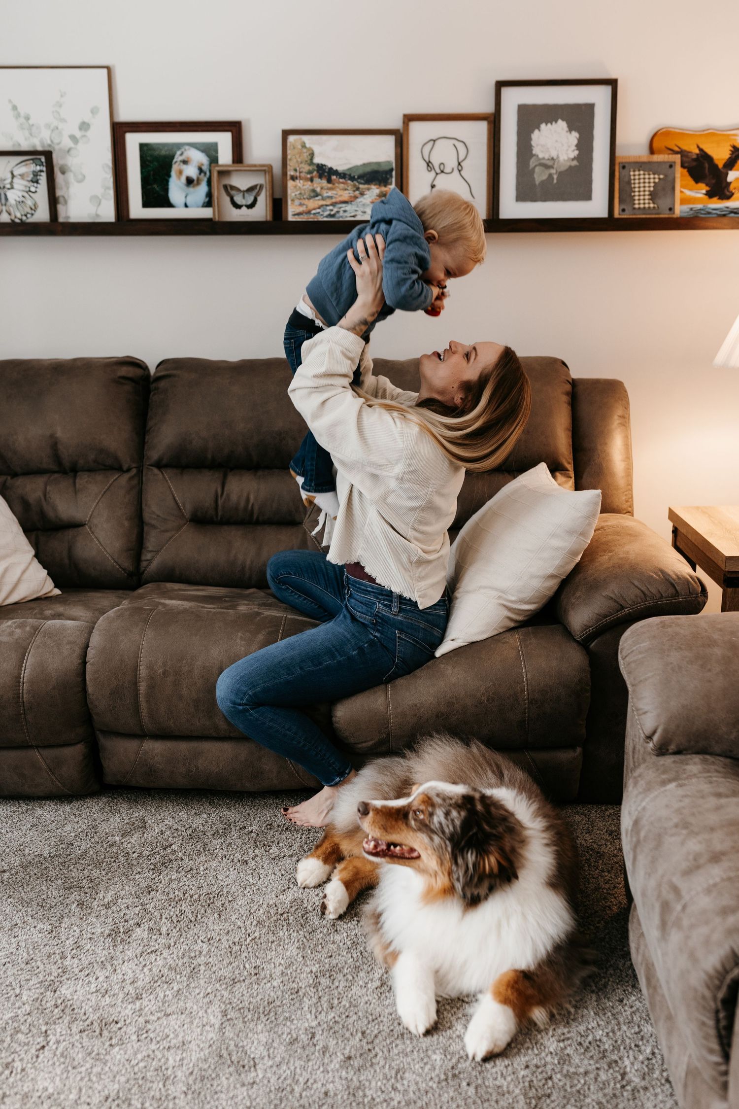 A person playing with a baby on a brown couch while a Shetland Sheepdog relaxes on the floor beside them.