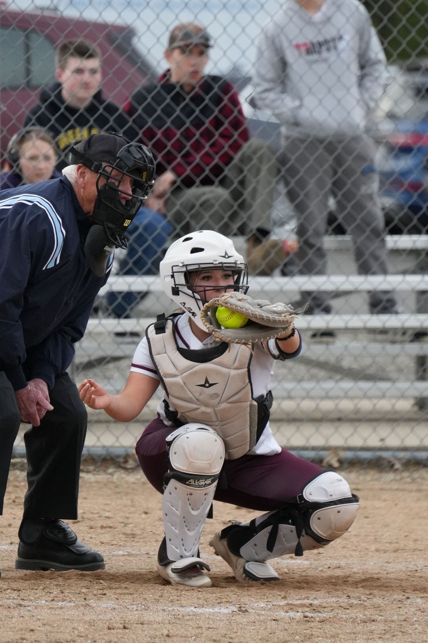 Female softball catcher in white helmet and gear crouches to catch a yellow ball, with umpire standing behind her.
