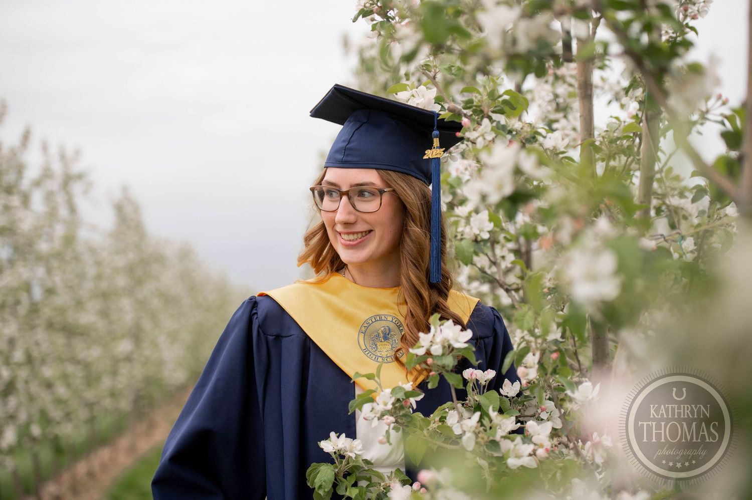Graduate in blue cap and gown poses among blooming apple trees while holding a bouquet and smiling.