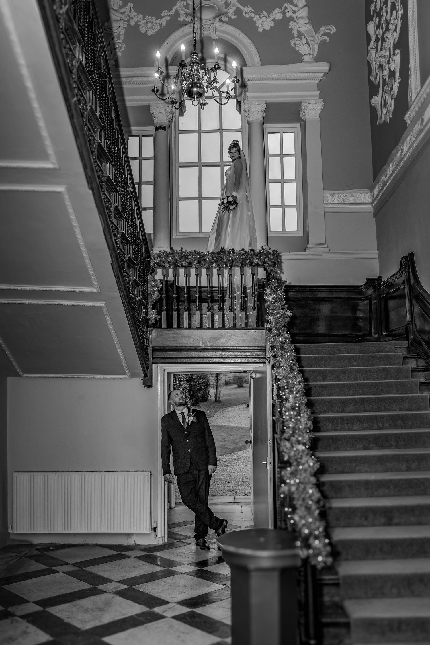 Artistic black and white photograph of an elegant interior staircase with chandelier and garland details.