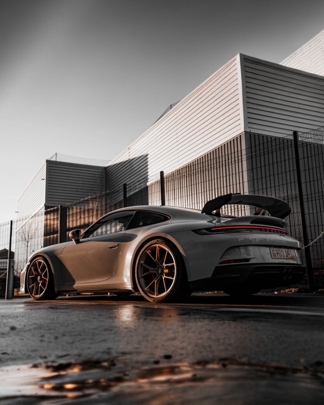Gray sports car with bronze wheels parked in front of modern industrial building on wet pavement.