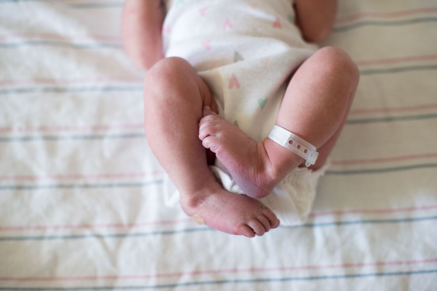 Small baby feet with hospital ID bracelet resting on white striped bedding.  Fresno Fresh 48 Hospital Photographer