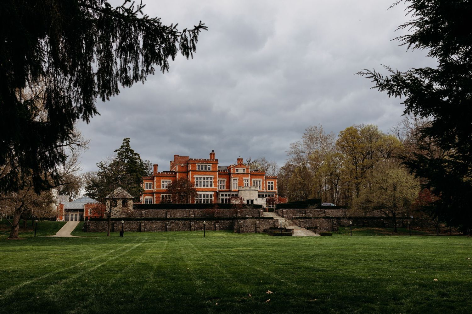 A grand red brick mansion with ornate architecture sits on manicured grounds under moody autumn skies.