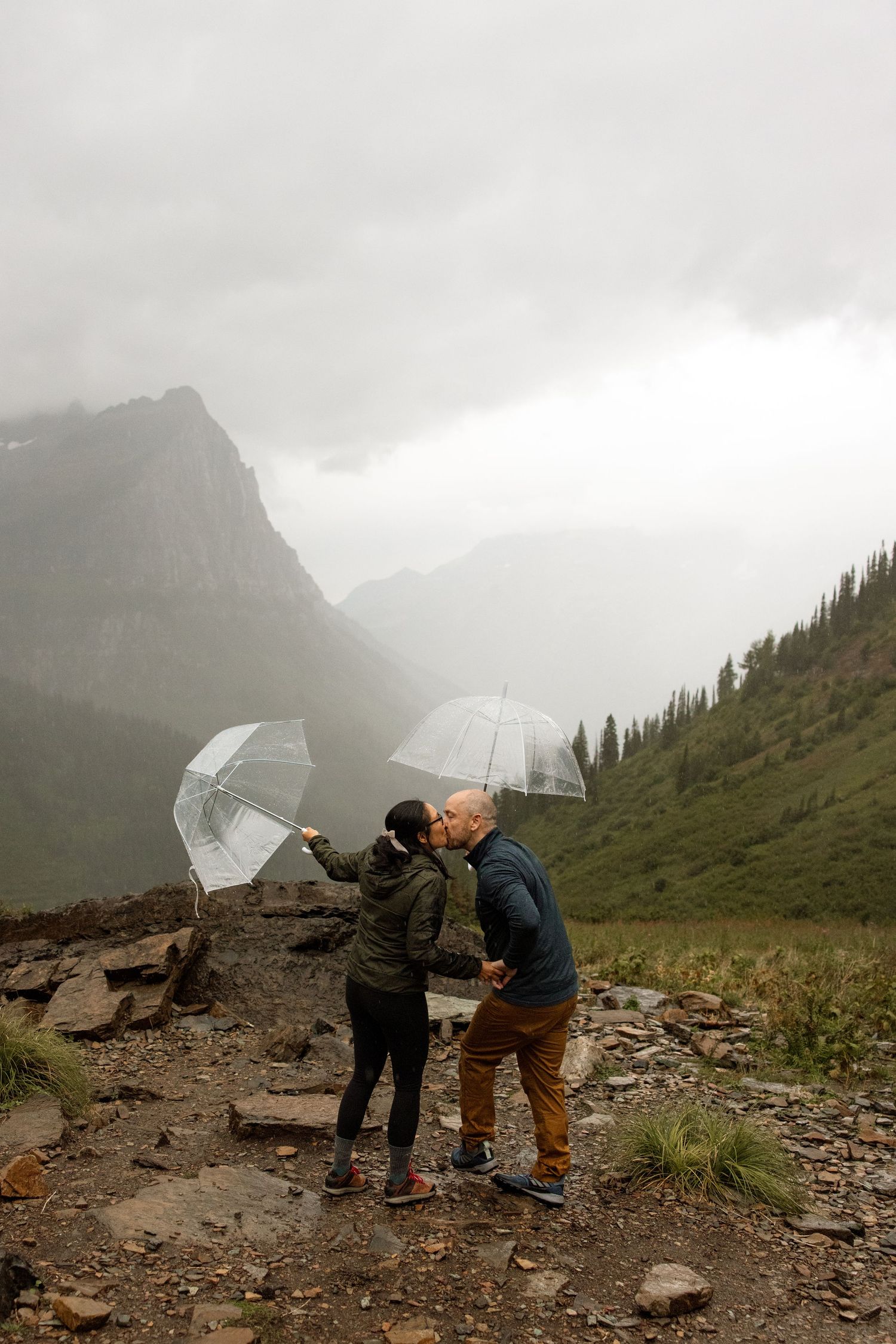 Two people with clear umbrellas share a romantic moment on a misty mountain trail in Glacier National Park.