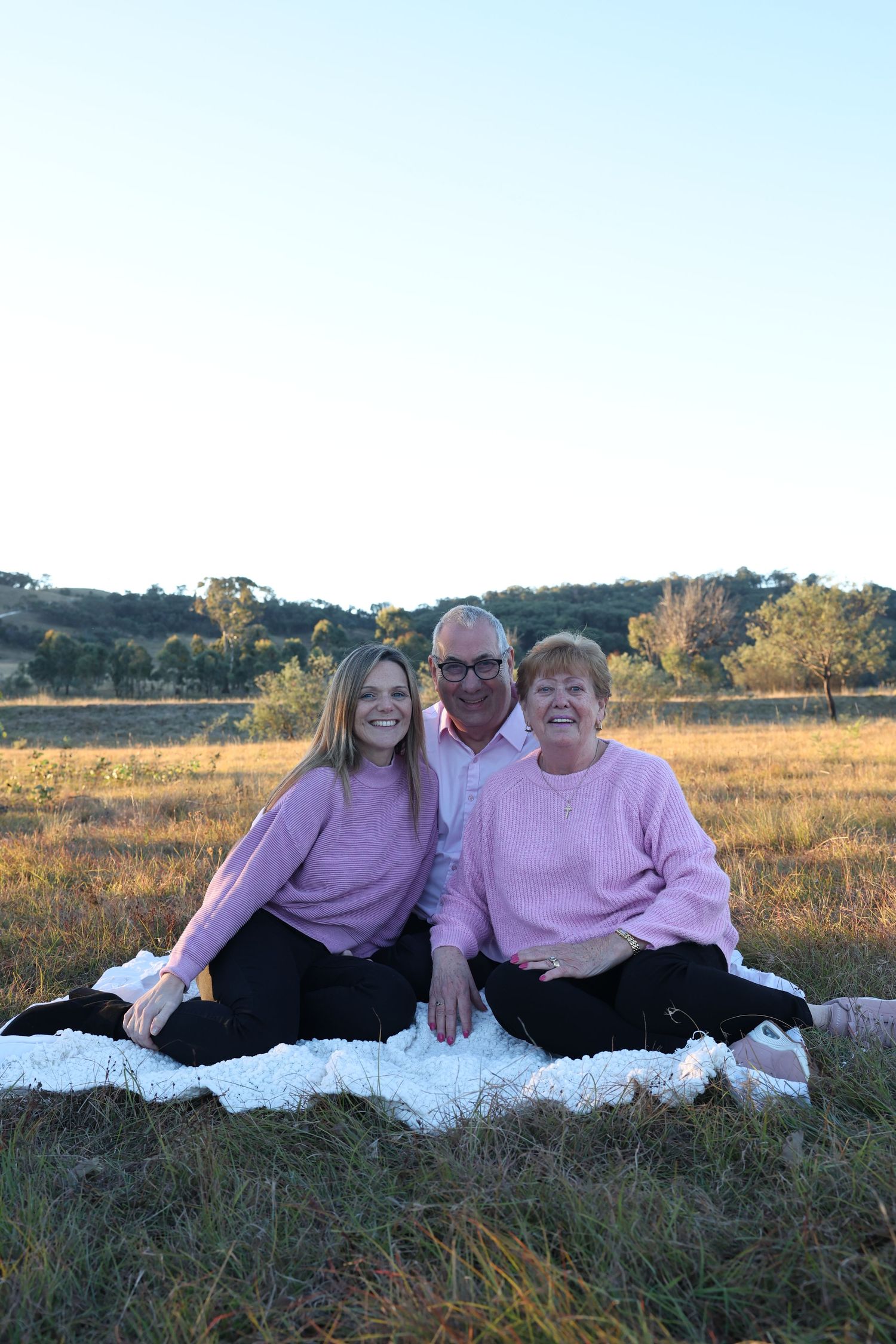 Three people wearing pink sweaters sit together on a white blanket in a golden grassy field at sunset.