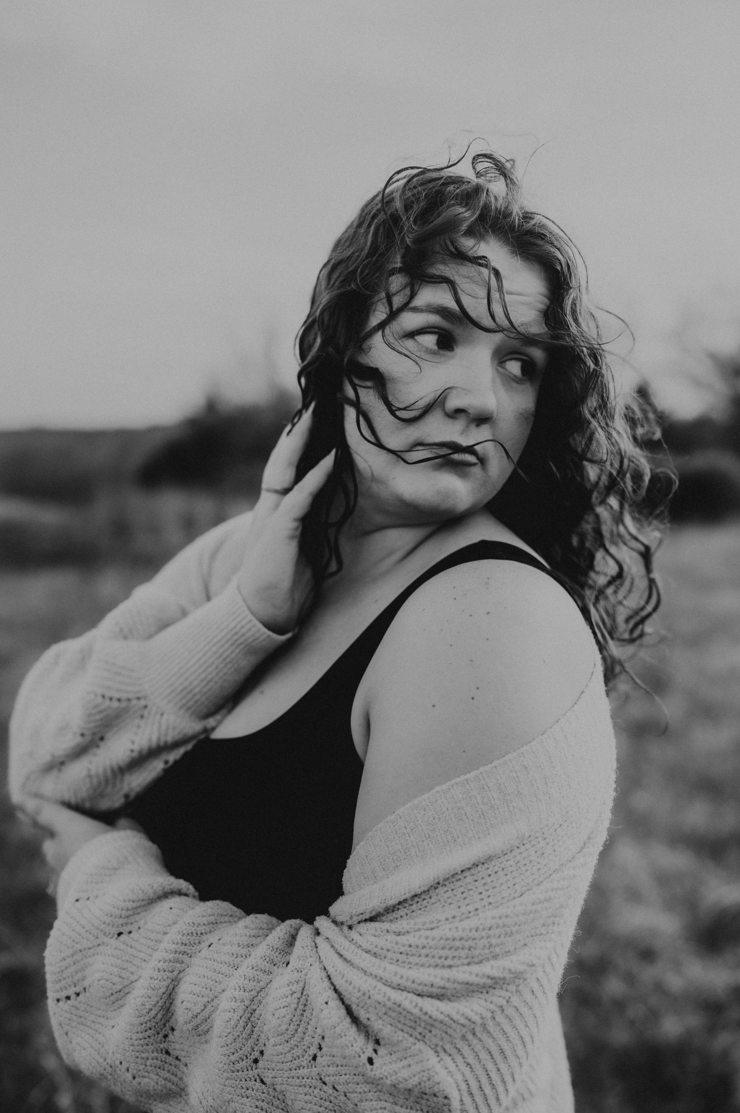 A black and white portrait of someone with curly hair wearing a cardigan and tank top in an outdoor setting.